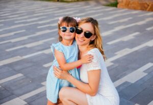 A woman in sunglasses kneels next to a young girl also wearing sunglasses as they embrace